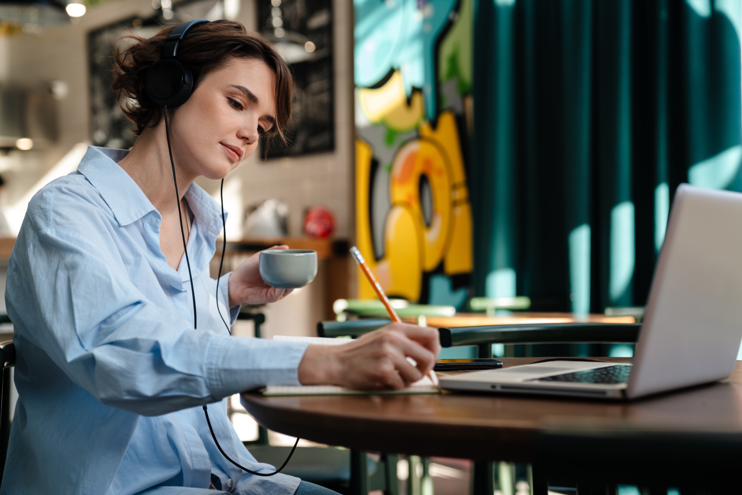 A woman with headphones holding a coffee and working calmly on a laptop in a coffee shop, illustrating how adults with ADHD can improve focus and productivity through monotasking instead of multitasking.